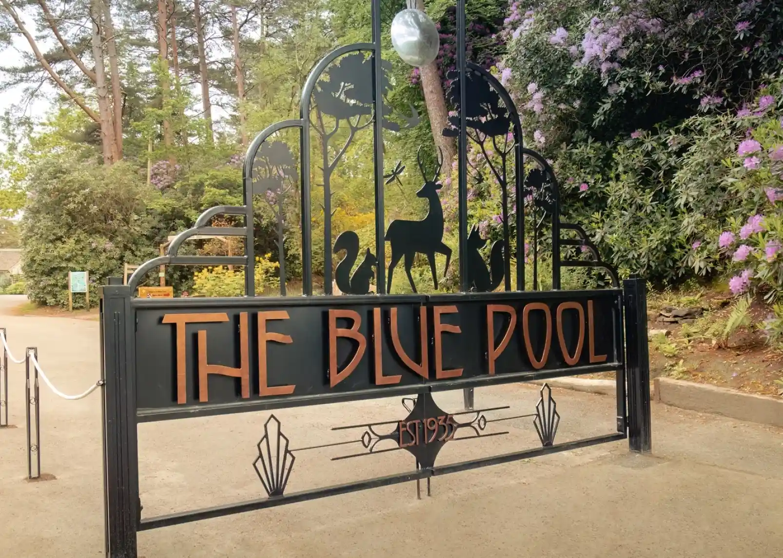 Decorative entrance gate to The Blue Pool, featuring artistic designs of animals and plants. "THE BLUE POOL" is prominently displayed, with "EST 1935" below. Surrounding greenery adds a natural backdrop.