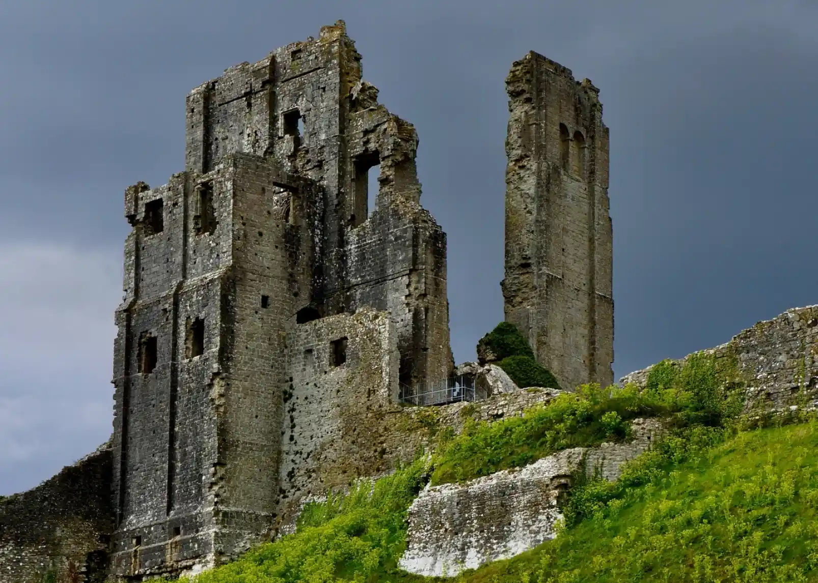 Medieval stone ruins of a castle stand on a grassy hill, partially covered in vegetation, under a dramatic cloudy sky.