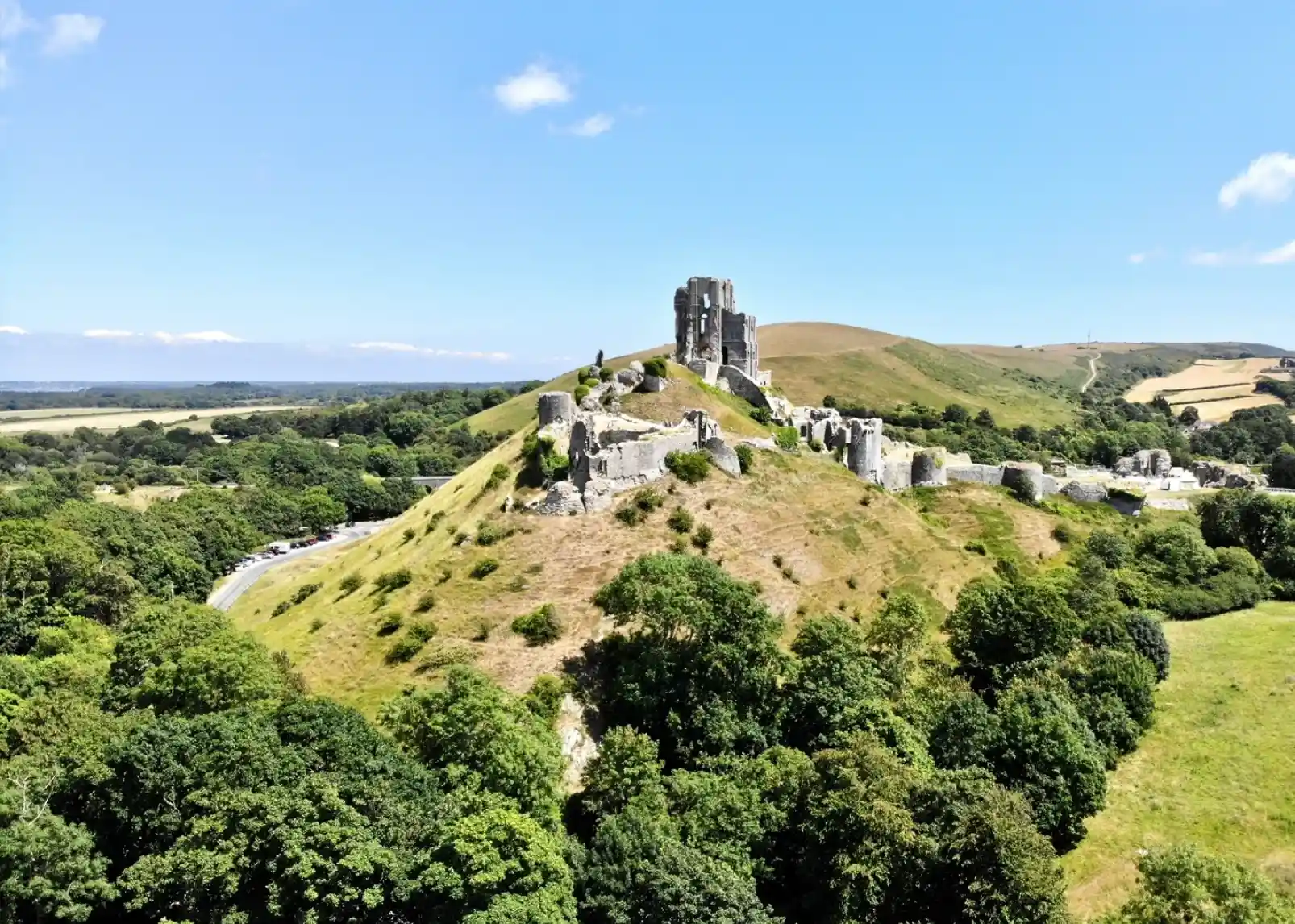 Aerial view of medieval ruins on a hillside, surrounded by lush greenery and rolling hills under a clear blue sky.