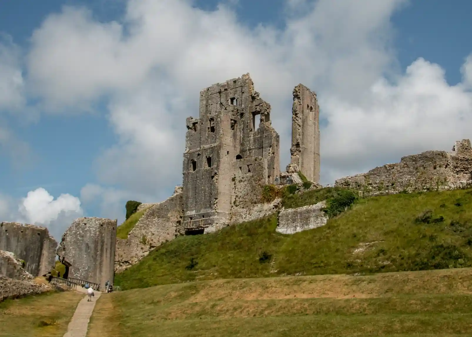 Medieval ruins of a stone castle on a grassy hill under a blue sky with scattered clouds. A pathway leads up to the remains, showcasing crumbling walls and towers.