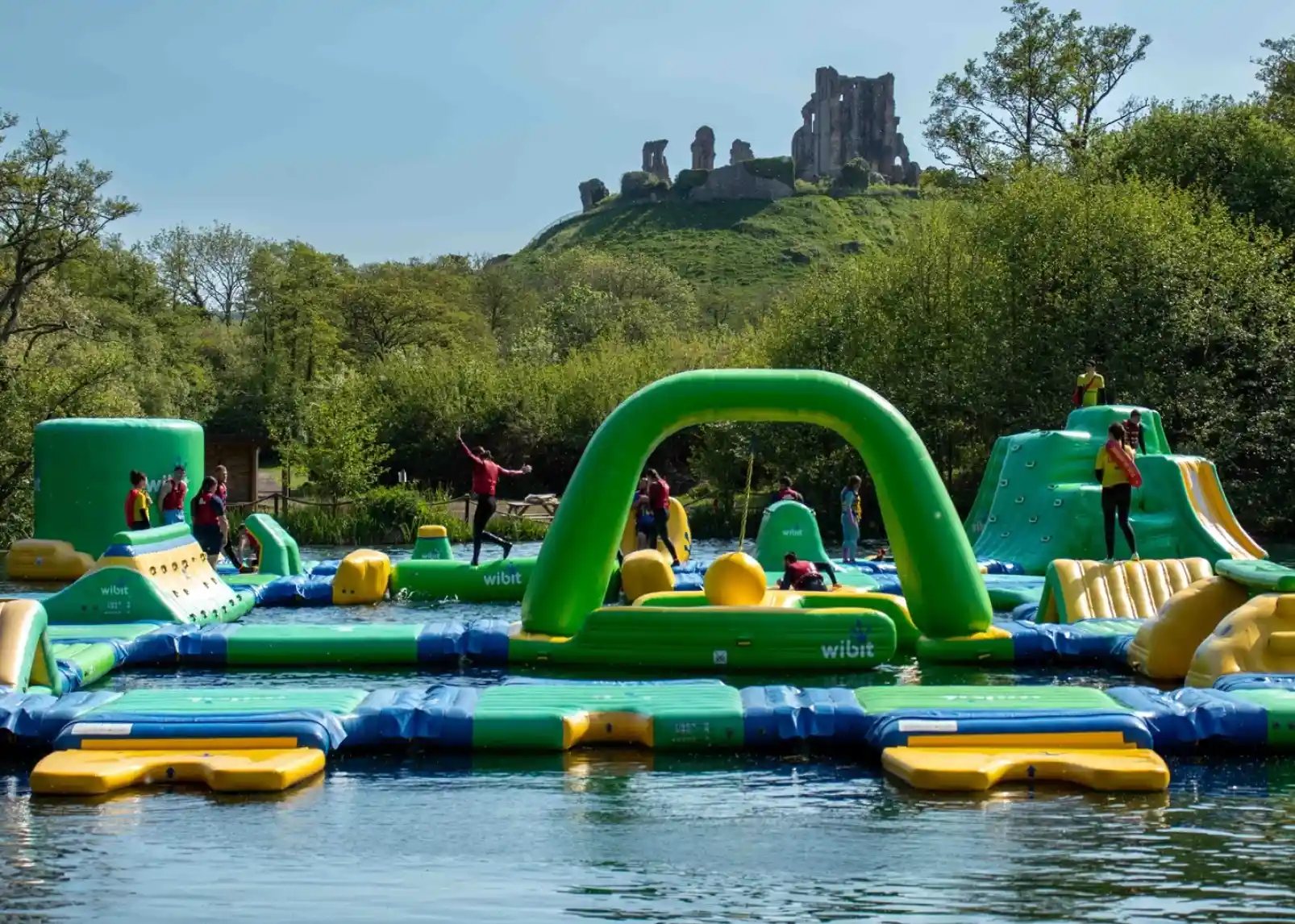 A vibrant inflatable water park is set up on a lake, with people actively engaging in various activities on the structures. In the background, a rocky hill topped with ruins is visible, surrounded by lush greenery under a clear blue sky.