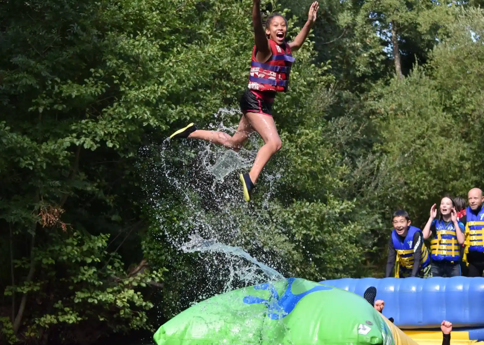 A vibrant inflatable water park is set up on a lake, with people actively engaging in various activities on the structures. In the background, a rocky hill topped with ruins is visible, surrounded by lush greenery under a clear blue sky.