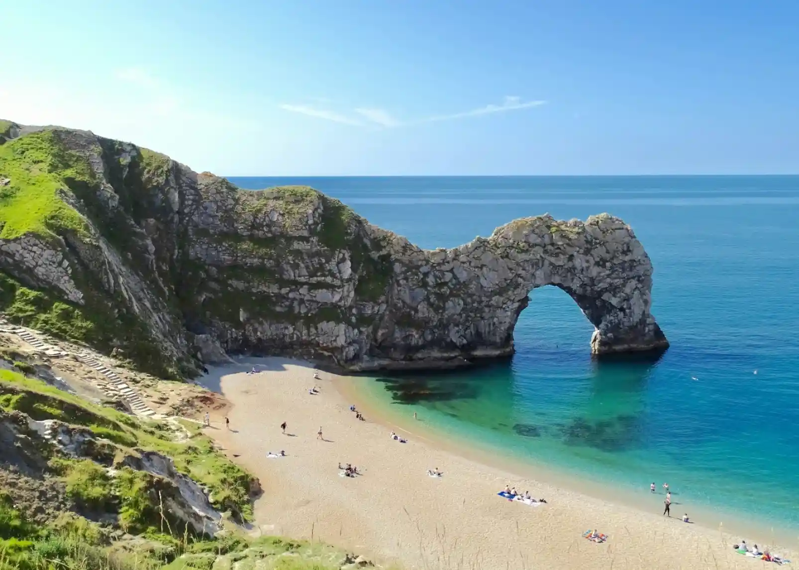 A scenic view of Durdle Door, a natural limestone arch, standing majestically over a sandy beach with turquoise waters. Lush green cliffs frame the scene under a clear blue sky. People sunbathe and stroll along the shore.