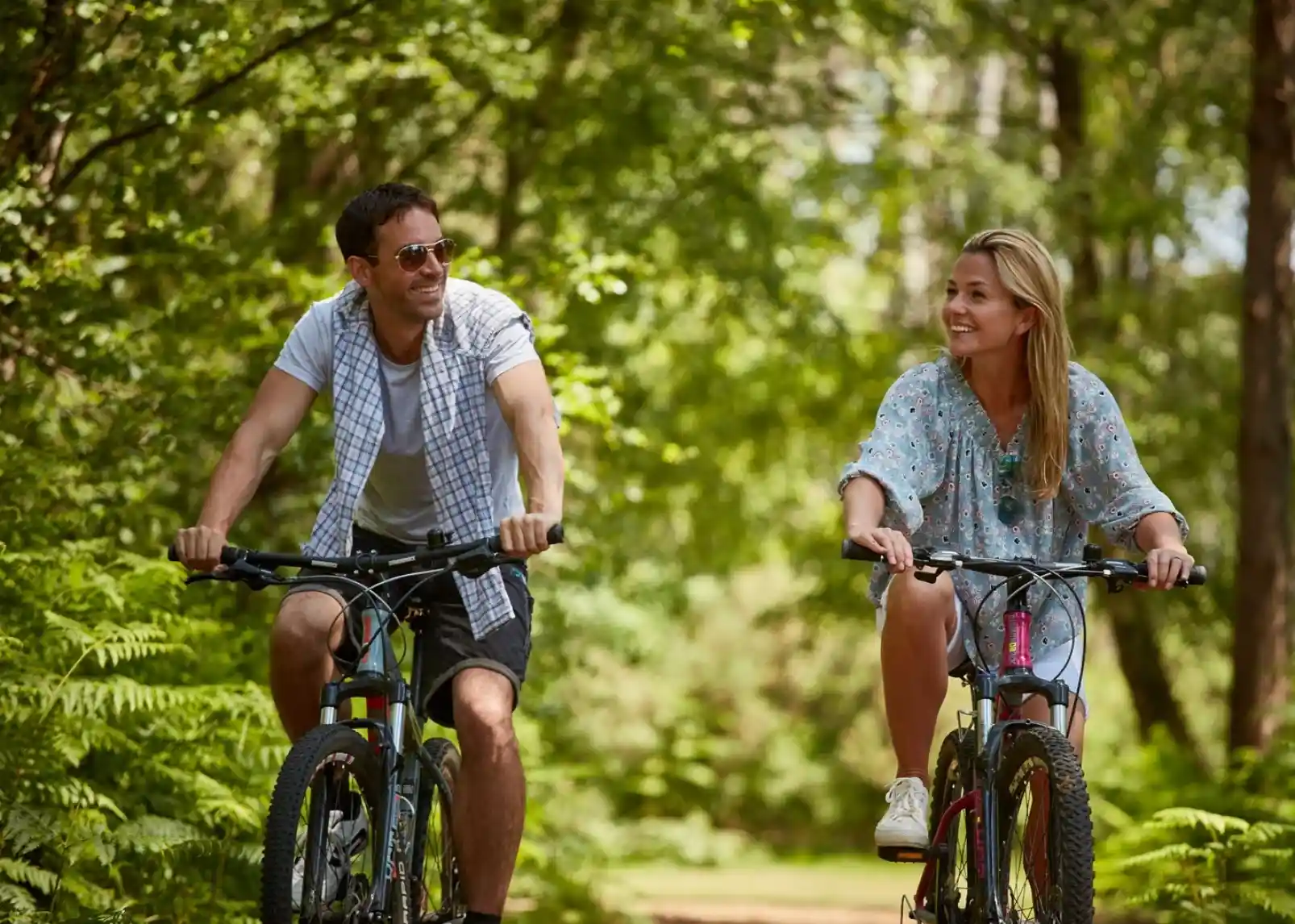 A man and woman ride bicycles along a wooded path, smiling at each other. Lush green foliage surrounds them, creating a serene outdoor atmosphere.