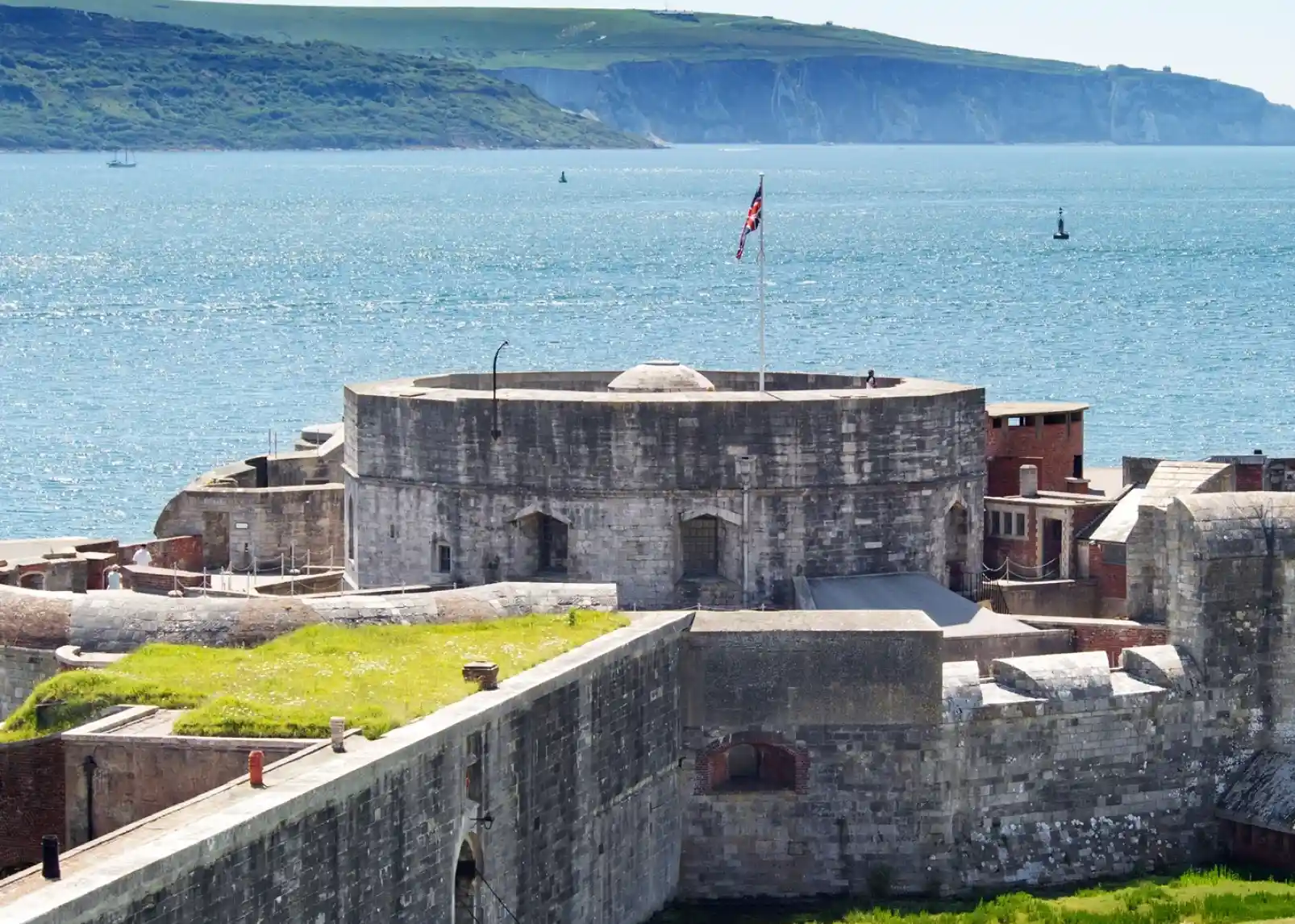 A coastal scene featuring a white lighthouse on a grassy shore, surrounded by a stone fortification and calm blue waters. Clear skies with scattered clouds and a distant green hillside add to the picturesque landscape.