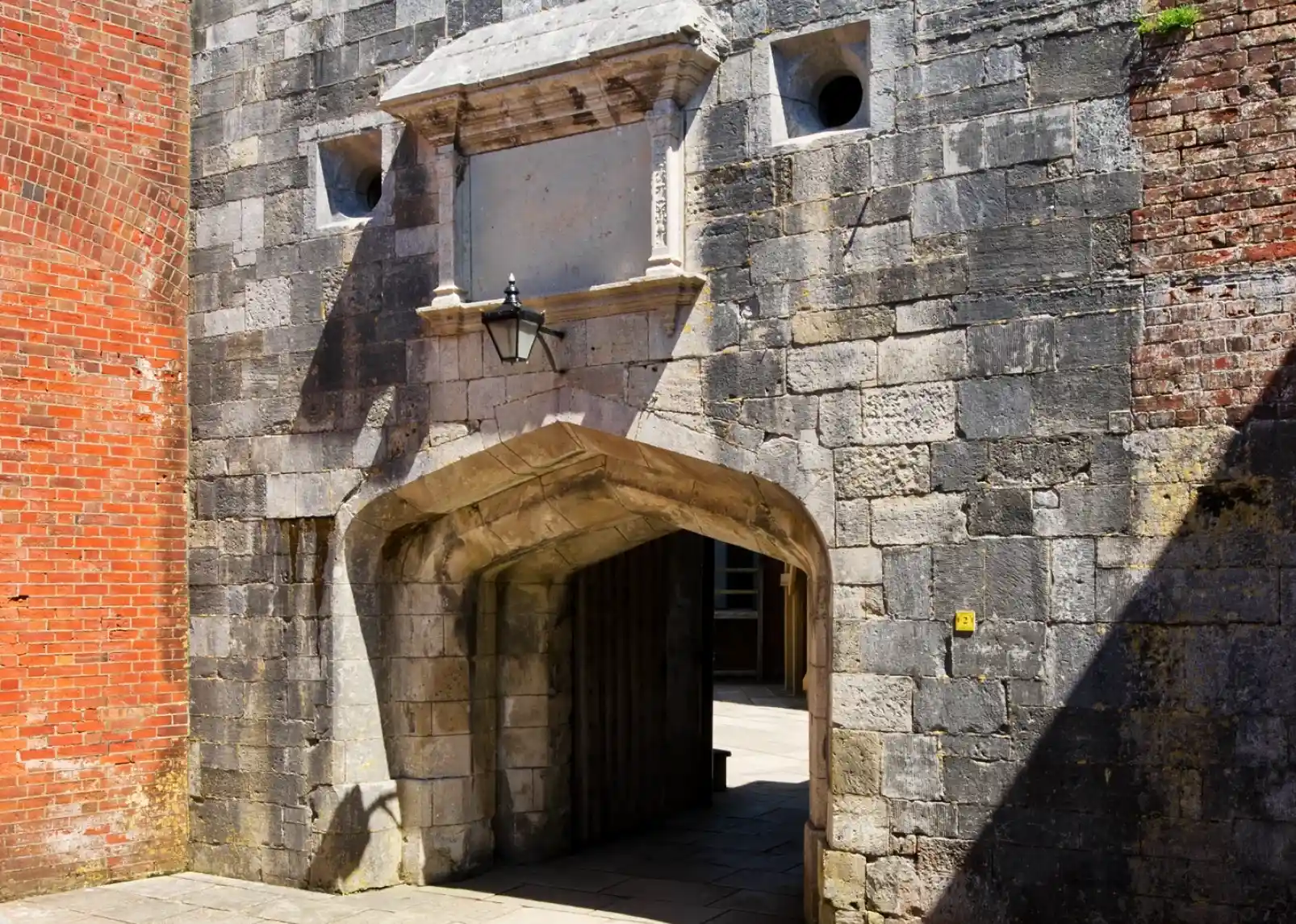 A coastal scene featuring a white lighthouse on a grassy shore, surrounded by a stone fortification and calm blue waters. Clear skies with scattered clouds and a distant green hillside add to the picturesque landscape.