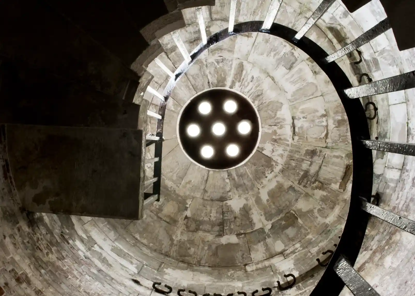 A coastal scene featuring a white lighthouse on a grassy shore, surrounded by a stone fortification and calm blue waters. Clear skies with scattered clouds and a distant green hillside add to the picturesque landscape.