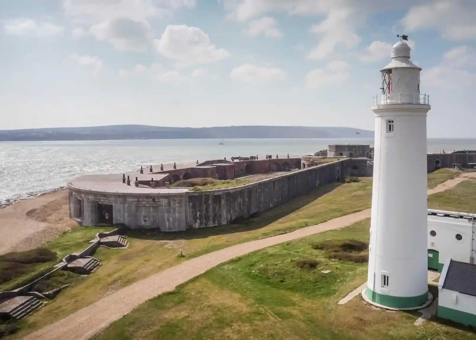 A coastal scene featuring a white lighthouse on a grassy shore, surrounded by a stone fortification and calm blue waters. Clear skies with scattered clouds and a distant green hillside add to the picturesque landscape.