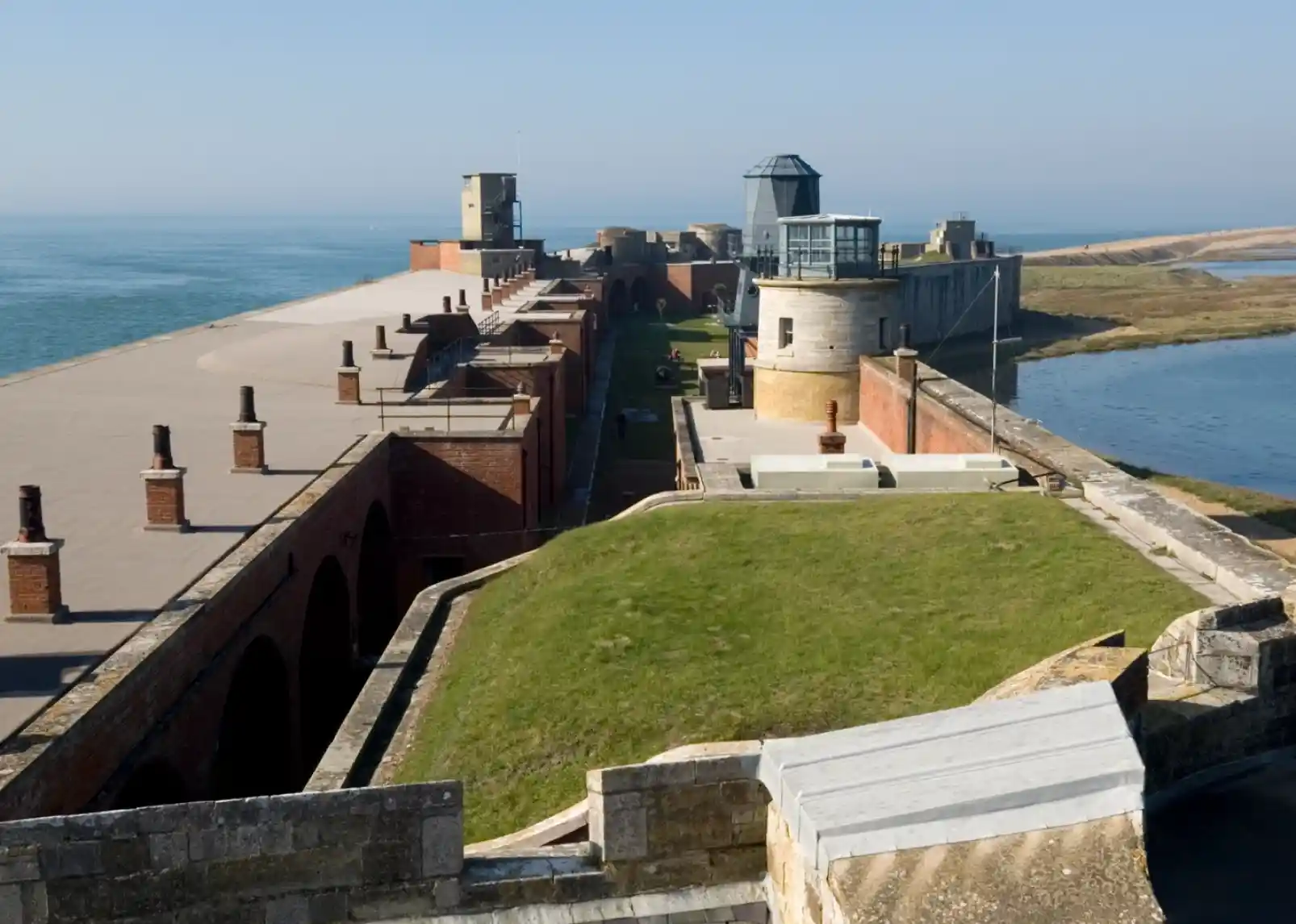 A coastal scene featuring a white lighthouse on a grassy shore, surrounded by a stone fortification and calm blue waters. Clear skies with scattered clouds and a distant green hillside add to the picturesque landscape.