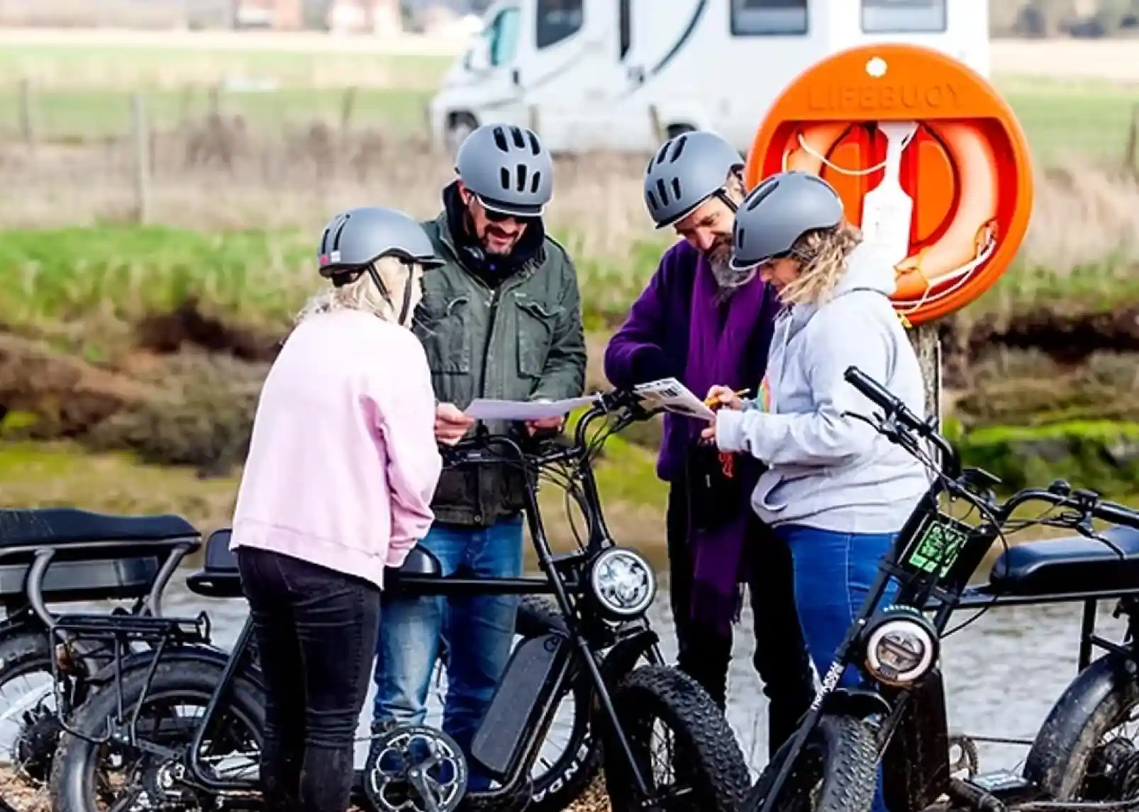 A group of seven people riding electric bikes on a gravel path, smiling and chatting. They are wearing helmets and casual clothing, with scenic hills in the background.