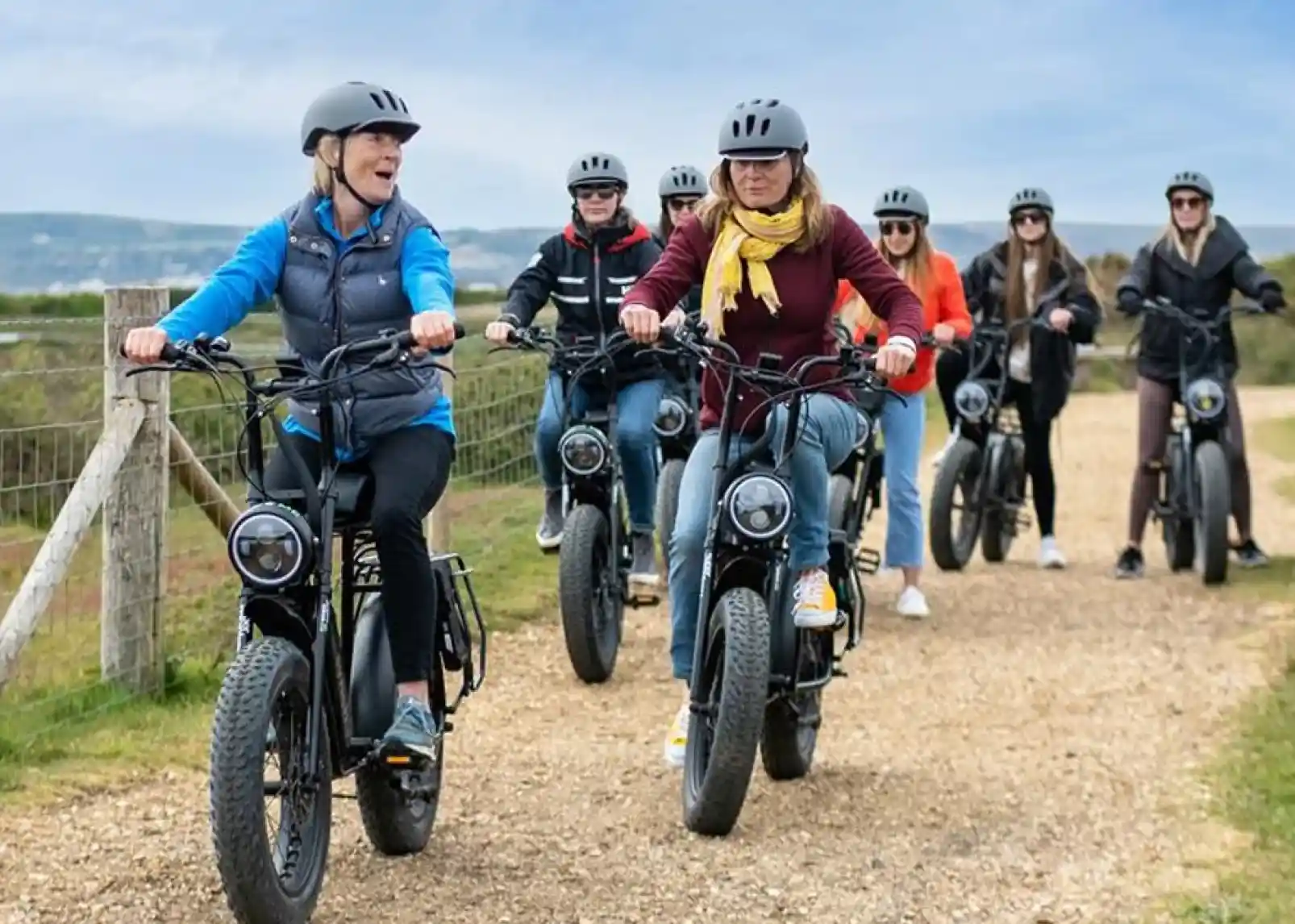 A group of seven people riding electric bikes on a gravel path, smiling and chatting. They are wearing helmets and casual clothing, with scenic hills in the background.