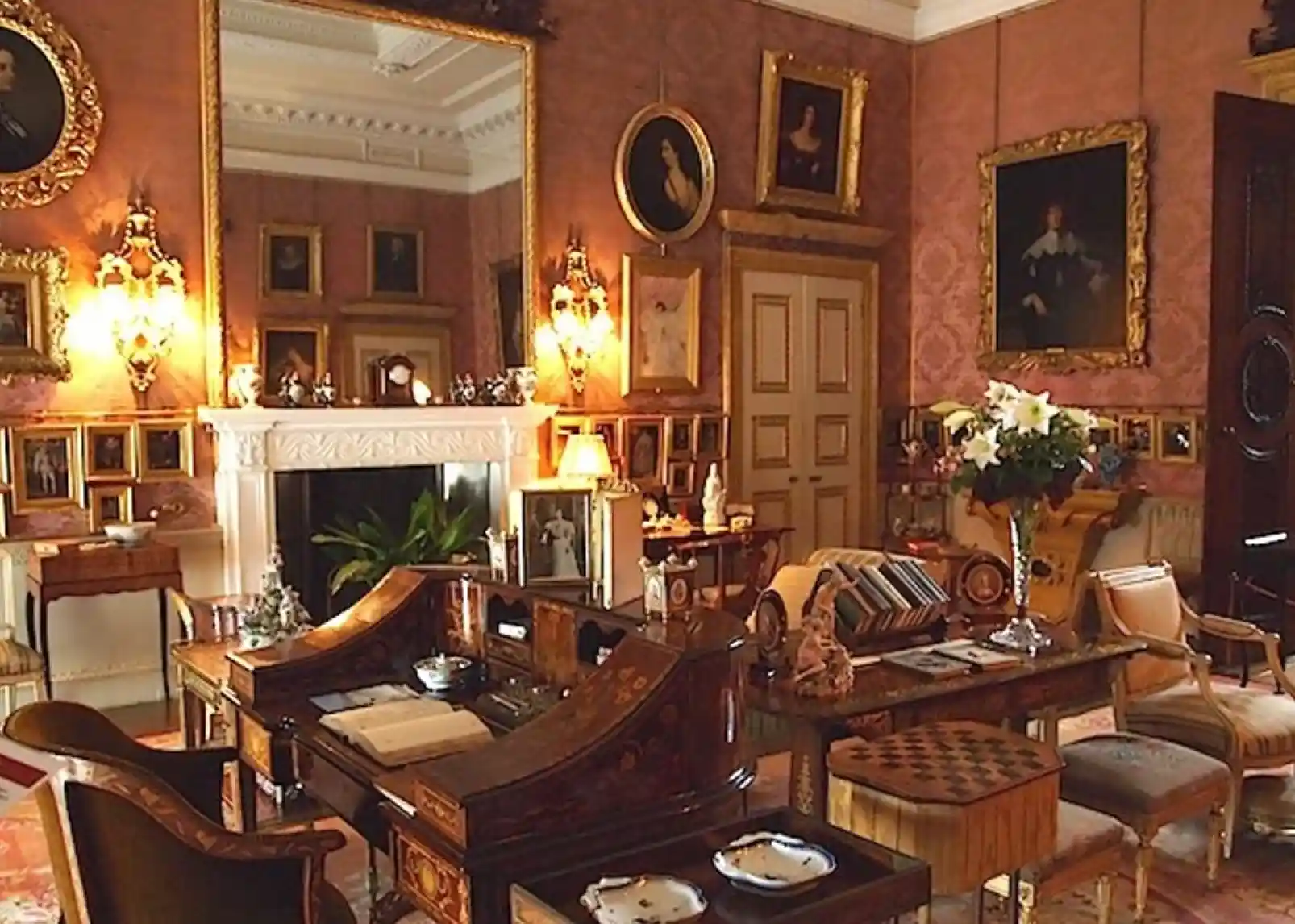 A grand, historic mansion with a slate roof and ornate facade is surrounded by neatly manicured gardens featuring colorful flower beds and sculpted hedges. Lush greenery and a wide pathway extend into the distance under a partly cloudy sky.