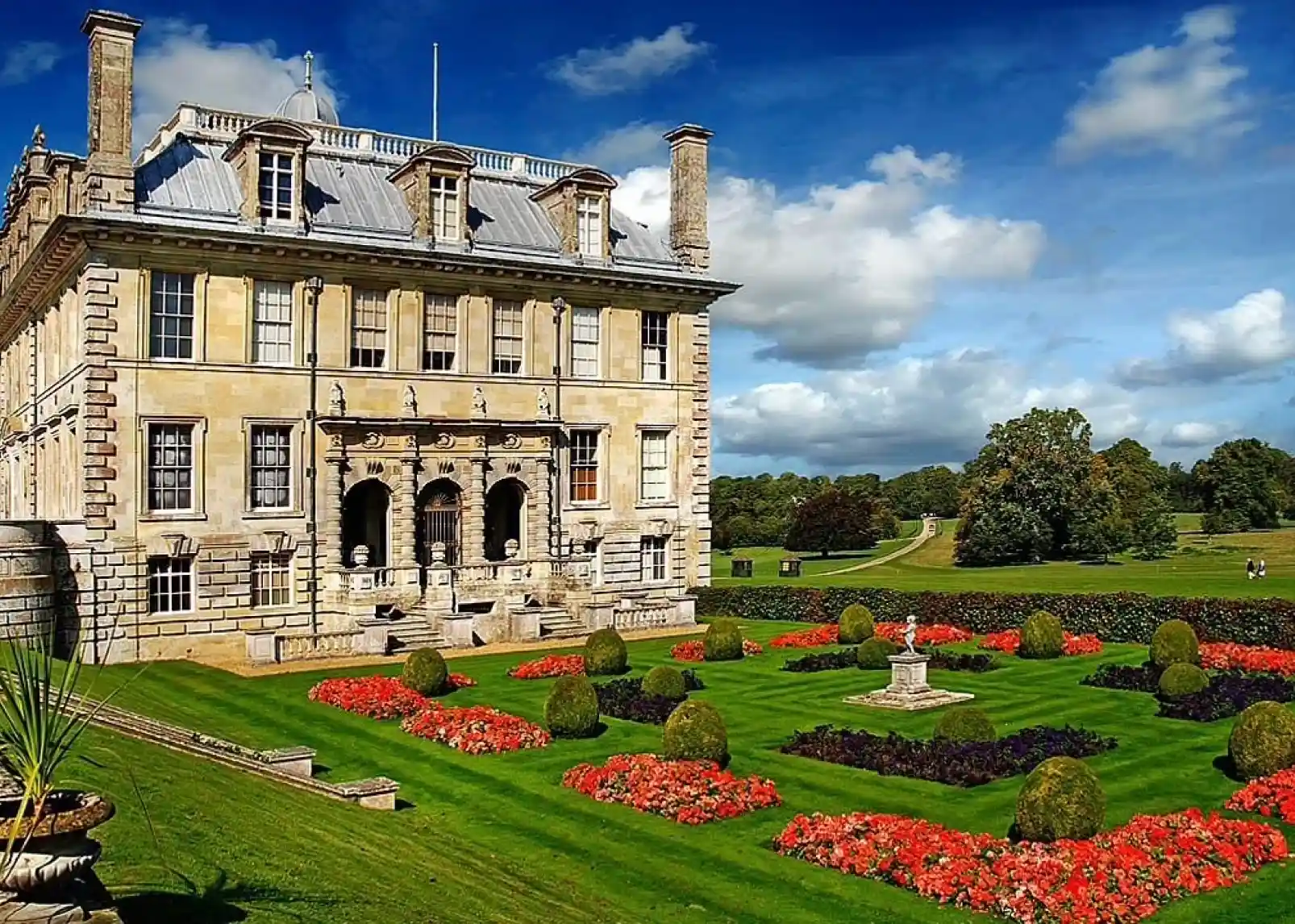 A grand, historic mansion with a slate roof and ornate facade is surrounded by neatly manicured gardens featuring colorful flower beds and sculpted hedges. Lush greenery and a wide pathway extend into the distance under a partly cloudy sky.