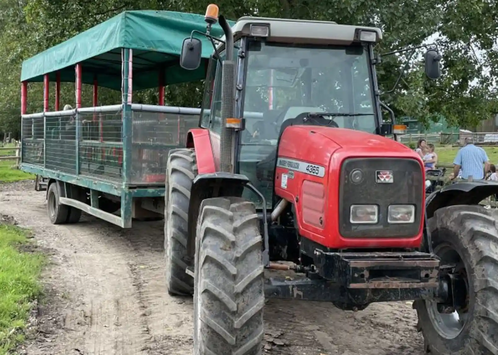 A smiling child with blonde hair holds a small, sleepy goat in her arms, both looking content. She is wearing a red sweatshirt, and a wooden fence is visible in the background.