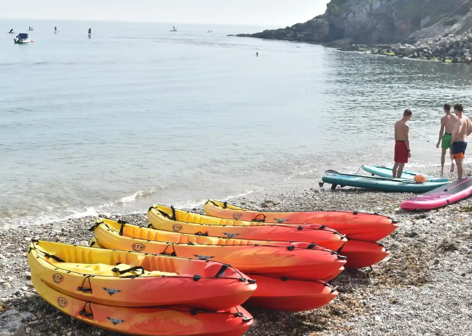 Several colorful kayaks are lined up on a rocky beach, while a few people stand nearby talking. In the background, various water activities take place, with paddleboarders and swimmers in the calm water. The shoreline is framed by a lush, green hillside.