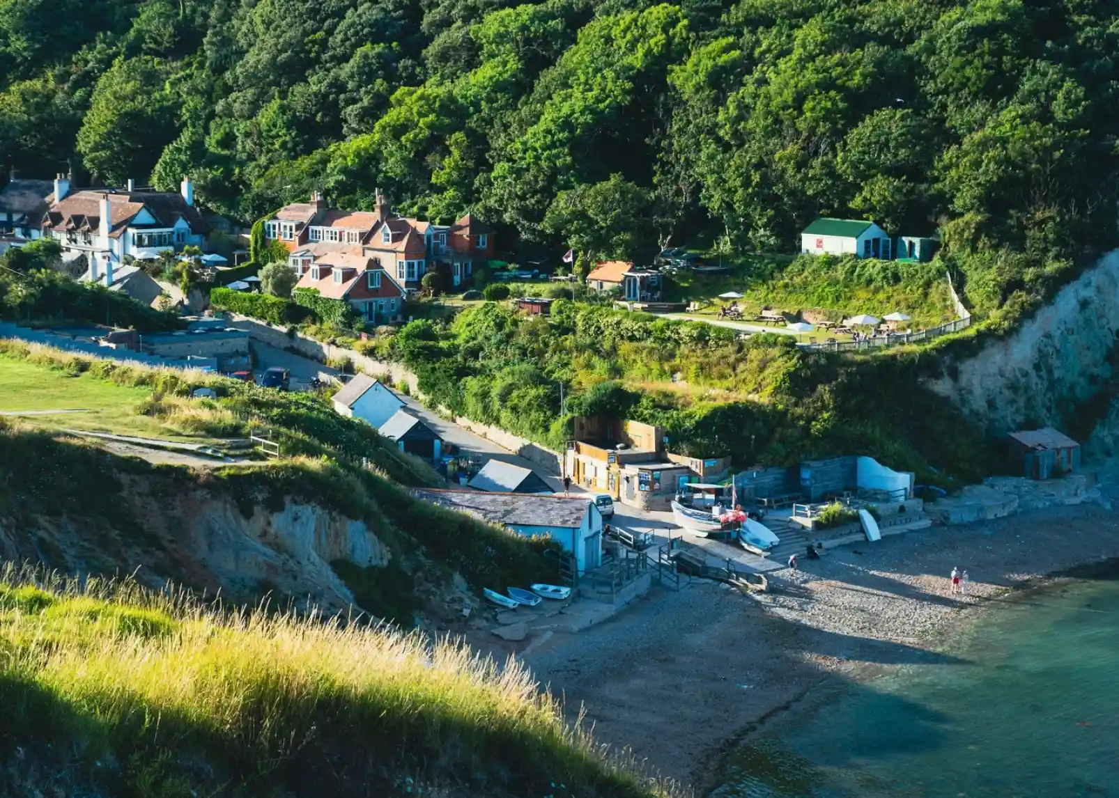 A scenic coastal view featuring a sandy beach with boats lined up, surrounded by greenery and hillside homes. The area has a mix of residential buildings and small structures, with people walking along the shore.