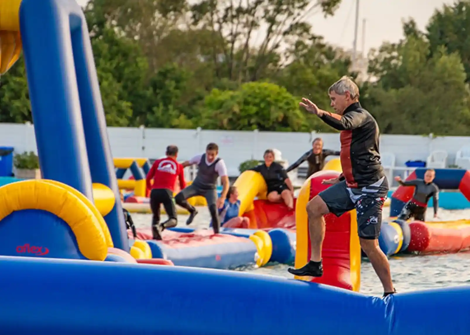 A man in a black shirt and shorts balances on a large inflatable structure at a water park. Others can be seen navigating various colorful inflatable obstacles in the background. Trees and a clear sky are visible, indicating a sunny day.