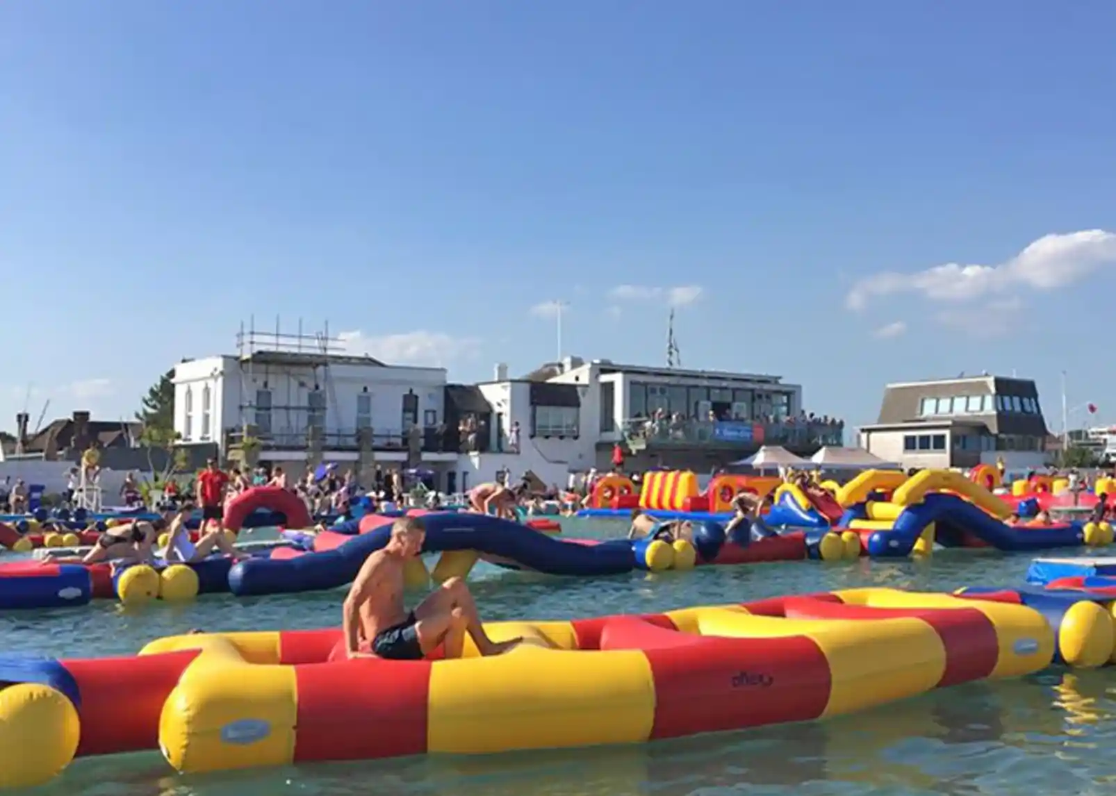 A colorful inflatable water park set in a lake, with various structures and people enjoying the activities. Bright blue sky and distant buildings provide a lively backdrop.