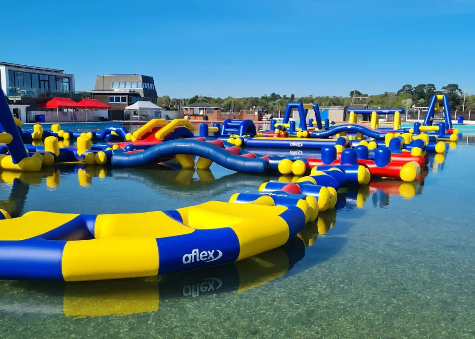 Colorful inflatable water park set up in a clear lake. The structure includes slides, bridges, and various obstacles, surrounded by trees and a blue sky. Sunlight reflects off the water.