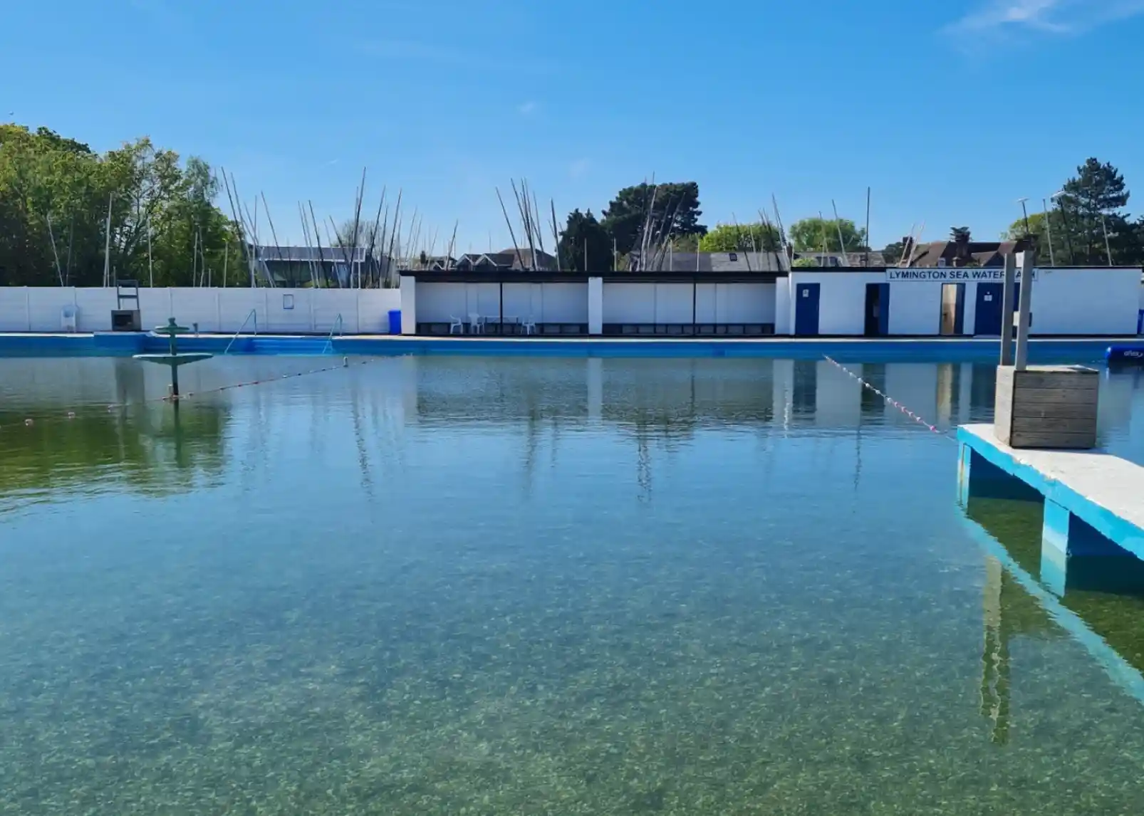 A calm outdoor swimming pool surrounded by greenery under a clear blue sky. Various sailboat masts are visible in the background, with buildings on one side of the pool. The water is clear, reflecting the surrounding environment.