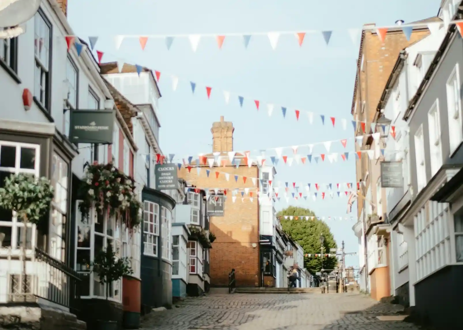 A quaint street lined with charming buildings, adorned with red, white, and blue bunting. Cobblestone path leads to a slightly elevated area with a tower in the distance.
