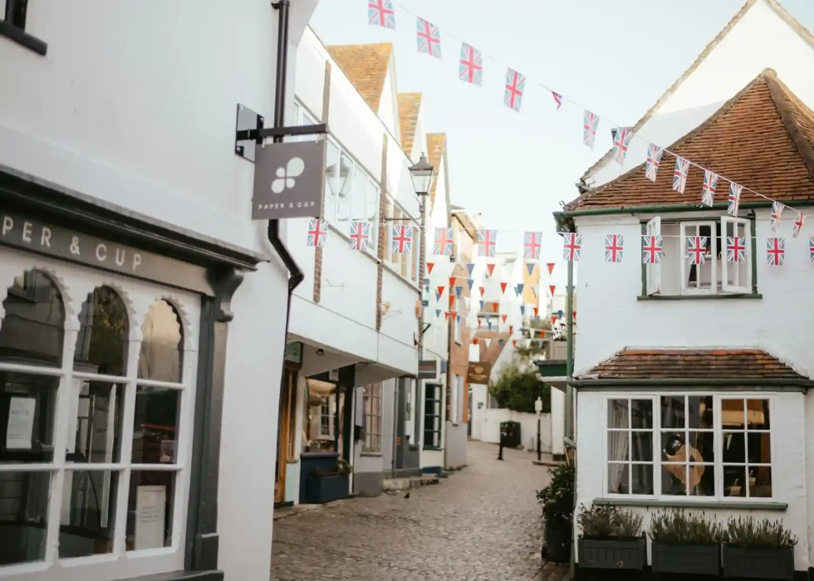 A quaint street lined with charming buildings, adorned with red, white, and blue bunting. Cobblestone path leads to a slightly elevated area with a tower in the distance.