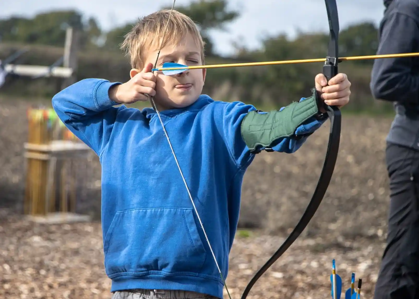 A family enjoys a sunny day canoeing on a calm river. Two adults and two children wear life jackets and are smiling while paddling. Lush greenery and reeds line the riverbank in the background.