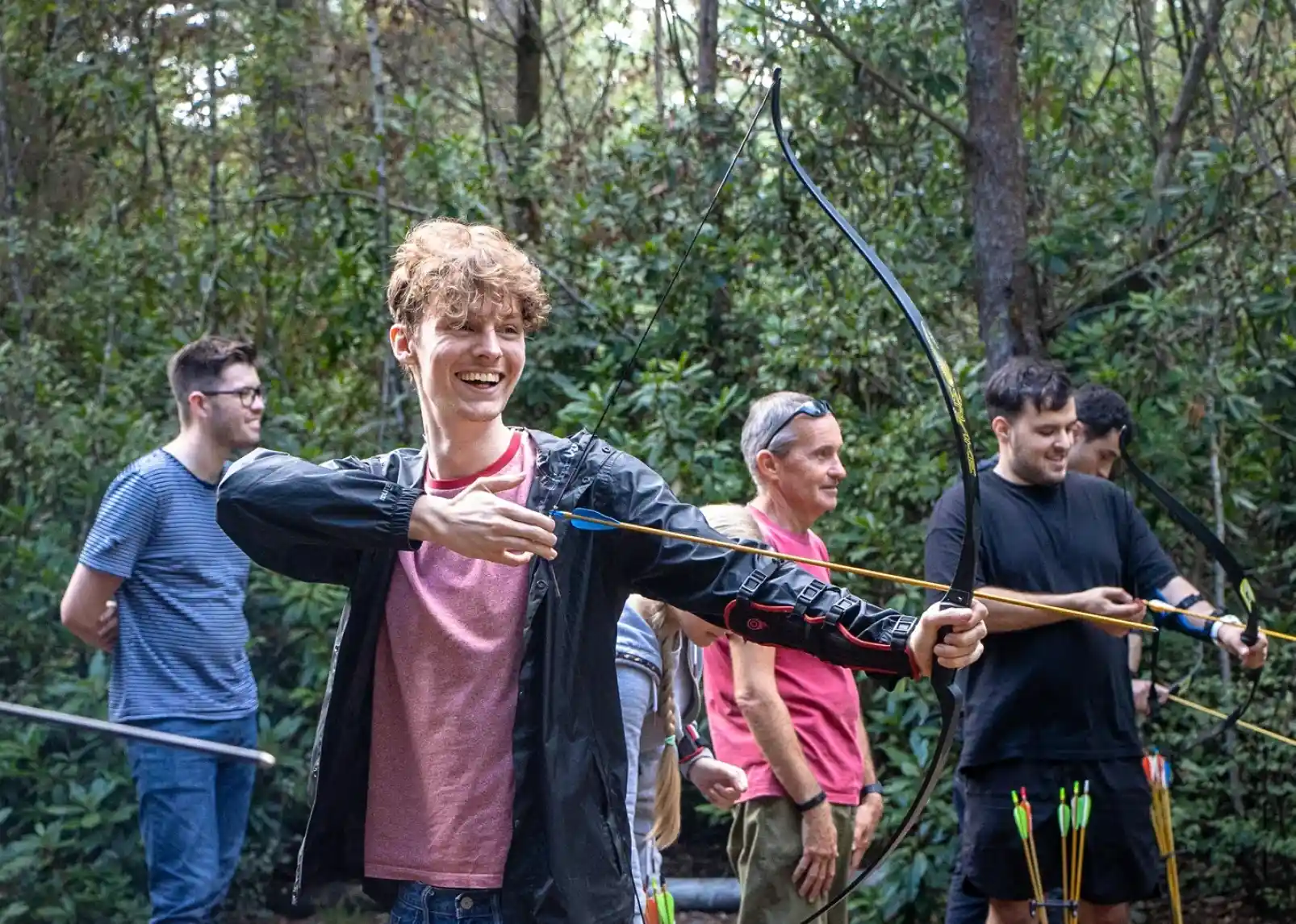 A family enjoys a sunny day canoeing on a calm river. Two adults and two children wear life jackets and are smiling while paddling. Lush greenery and reeds line the riverbank in the background.