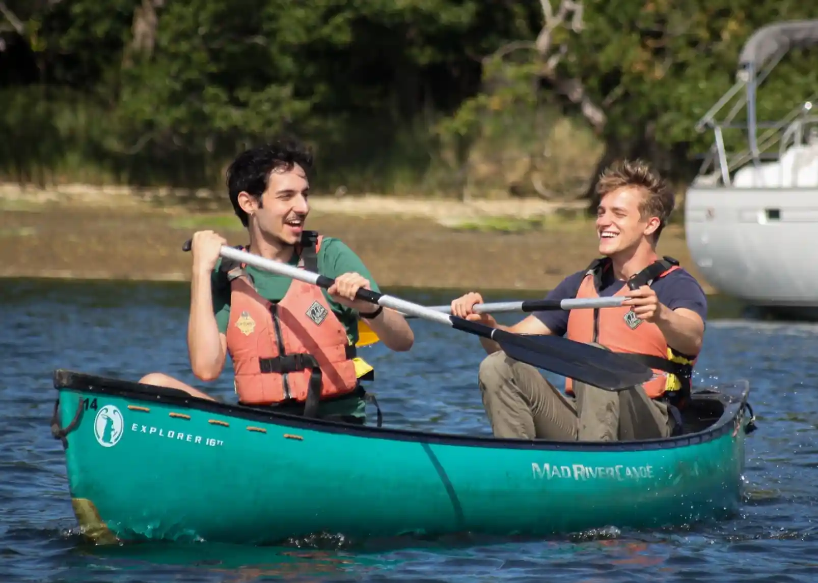 A family enjoys a sunny day canoeing on a calm river. Two adults and two children wear life jackets and are smiling while paddling. Lush greenery and reeds line the riverbank in the background.