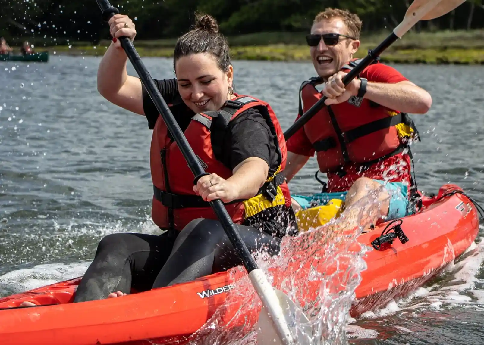 A family enjoys a sunny day canoeing on a calm river. Two adults and two children wear life jackets and are smiling while paddling. Lush greenery and reeds line the riverbank in the background.