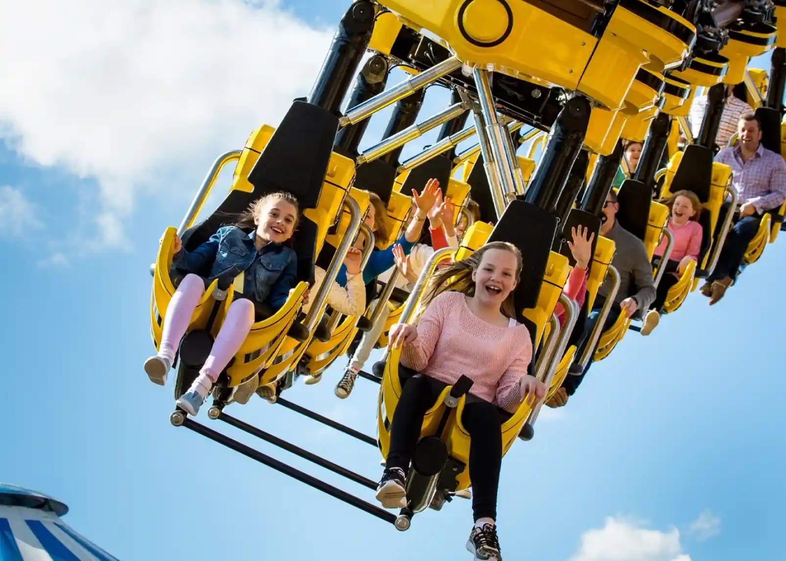 A roller coaster with vintage-style cars zooms through a loop under a clear blue sky. Riders are enjoying the thrill, with some raising their arms in excitement. The track is a bright yellow, contrasting with the surrounding landscape.