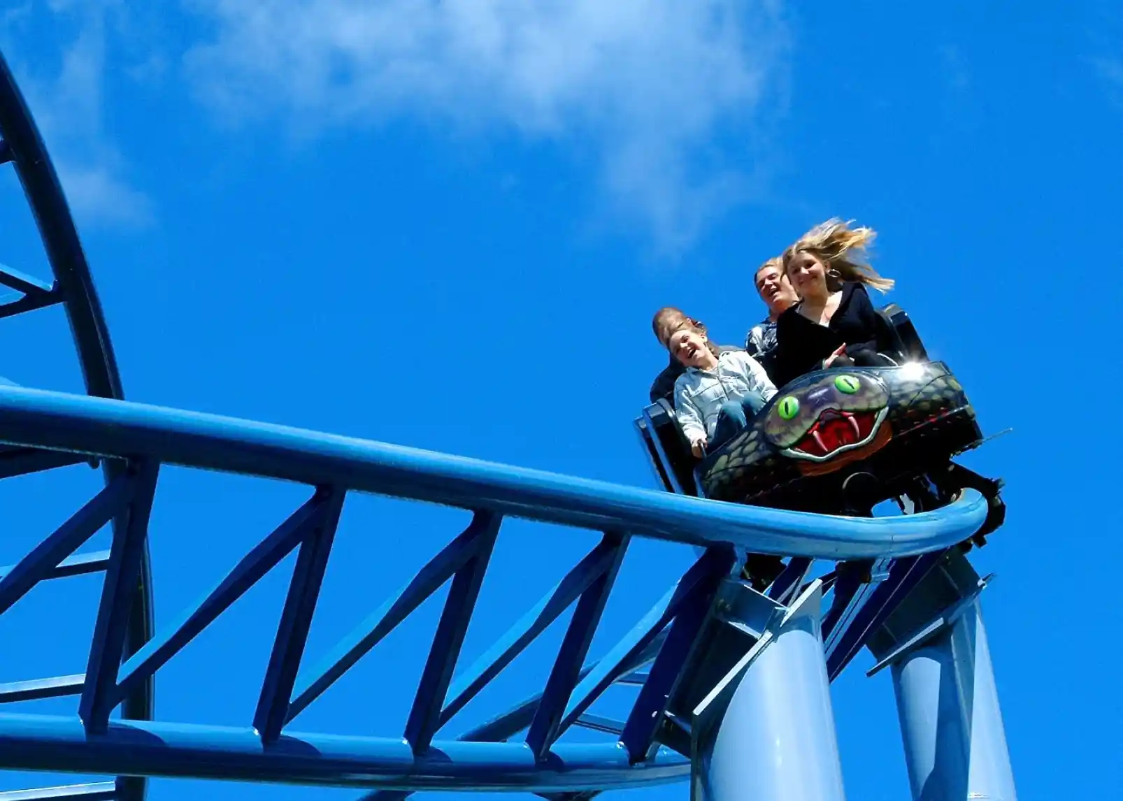 A roller coaster with vintage-style cars zooms through a loop under a clear blue sky. Riders are enjoying the thrill, with some raising their arms in excitement. The track is a bright yellow, contrasting with the surrounding landscape.