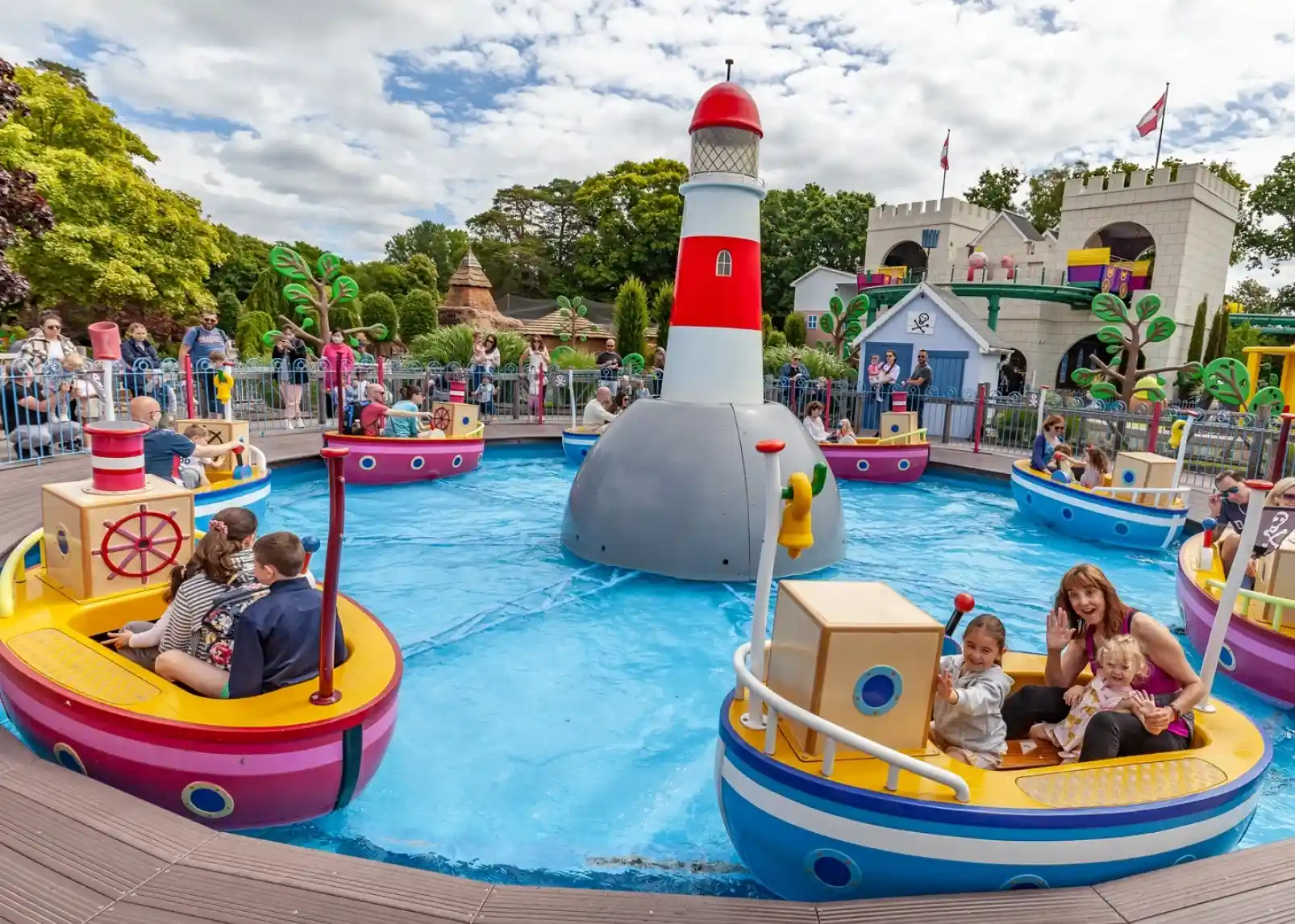 A group of animated characters from a children's show stands together, wearing colorful outfits. They are set against a backdrop of amusement park attractions, including colorful hot air balloons. The characters are smiling and appear cheerful.