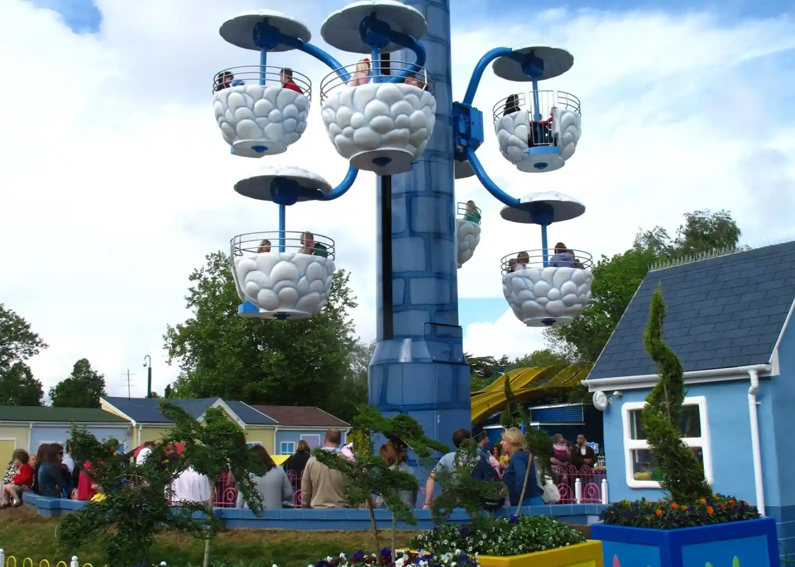A group of animated characters from a children's show stands together, wearing colorful outfits. They are set against a backdrop of amusement park attractions, including colorful hot air balloons. The characters are smiling and appear cheerful.