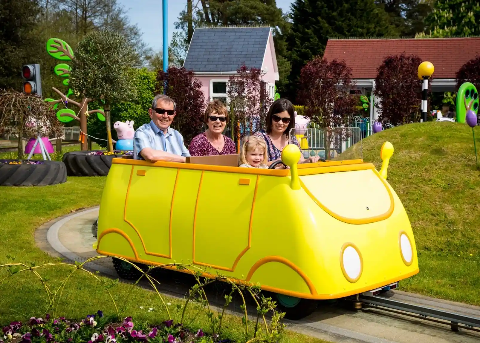 A group of animated characters from a children's show stands together, wearing colorful outfits. They are set against a backdrop of amusement park attractions, including colorful hot air balloons. The characters are smiling and appear cheerful.
