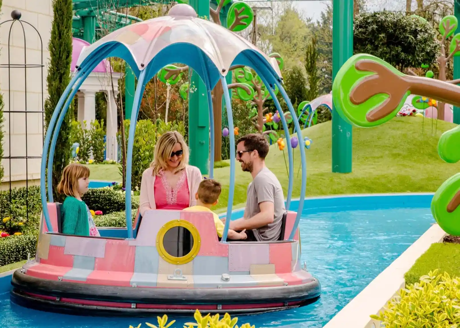 A group of animated characters from a children's show stands together, wearing colorful outfits. They are set against a backdrop of amusement park attractions, including colorful hot air balloons. The characters are smiling and appear cheerful.