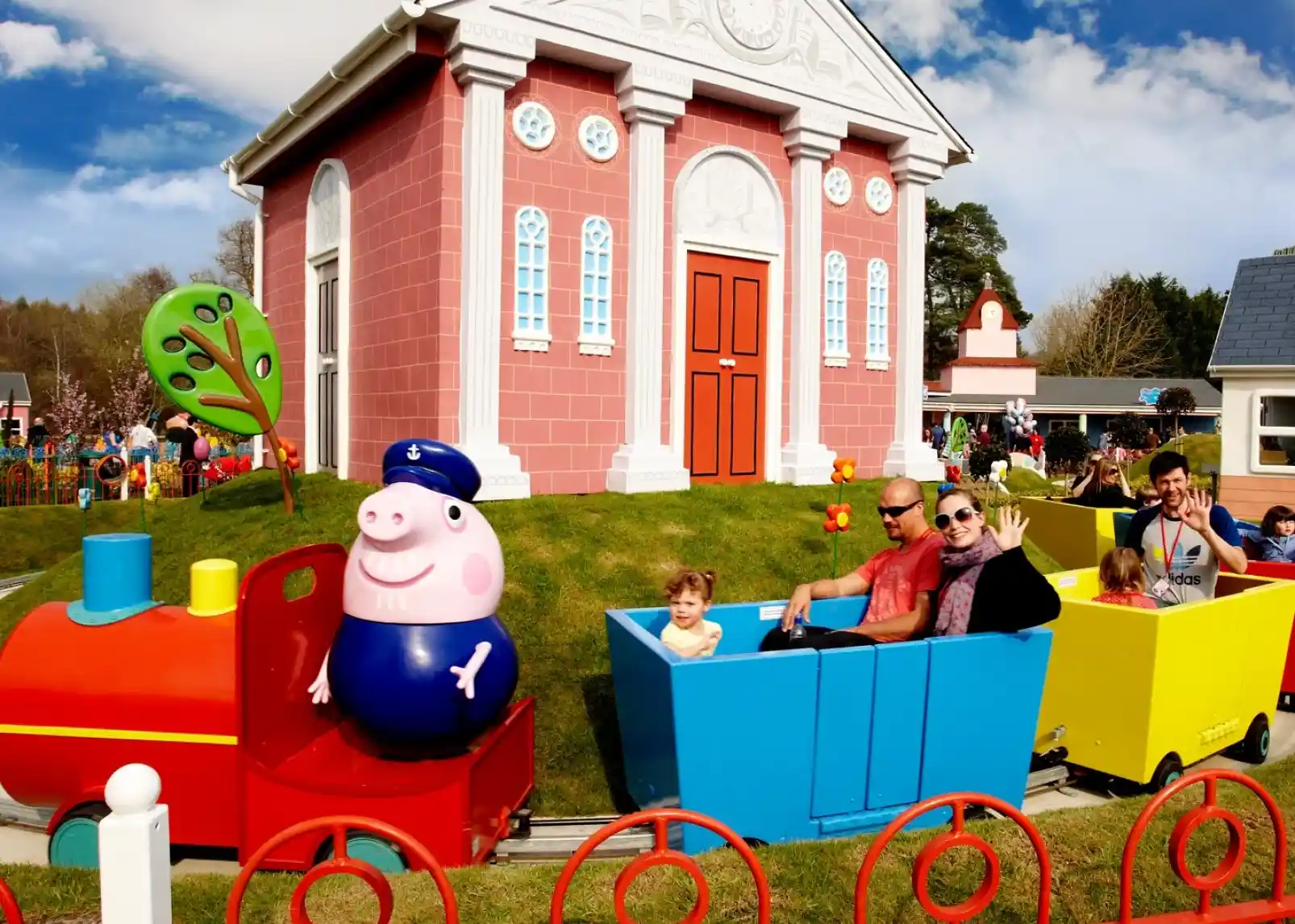 A group of animated characters from a children's show stands together, wearing colorful outfits. They are set against a backdrop of amusement park attractions, including colorful hot air balloons. The characters are smiling and appear cheerful.