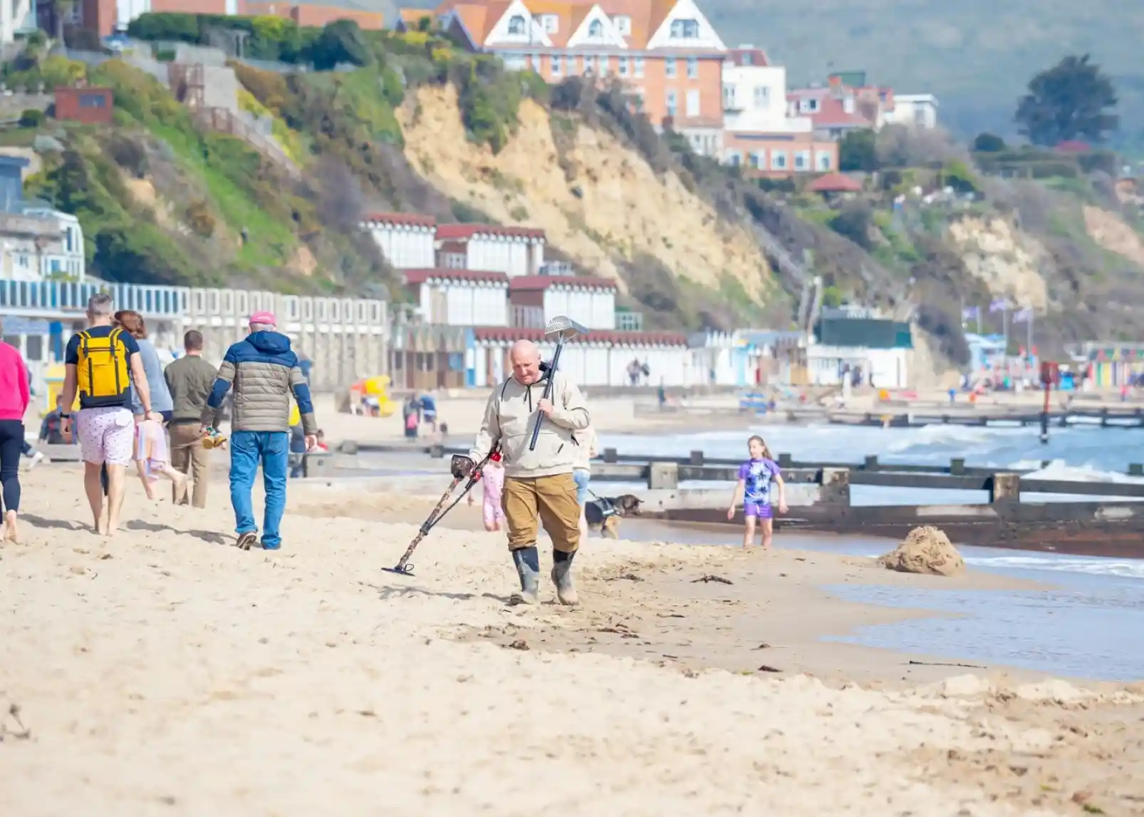 A man with a metal detector walks along a sandy beach, searching for items. People stroll in the background, while the coastline features cliffs and beach houses. Some children play near the shore, enjoying a sunny day.