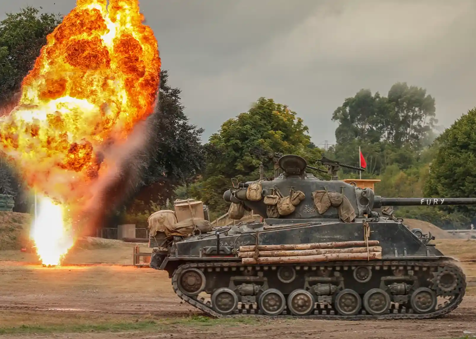 A military tank labeled "FURY" stands on a grassy field, with a large explosion of fire and debris erupting in the background. Thick smoke and orange flames contrast against a cloudy sky and green trees.