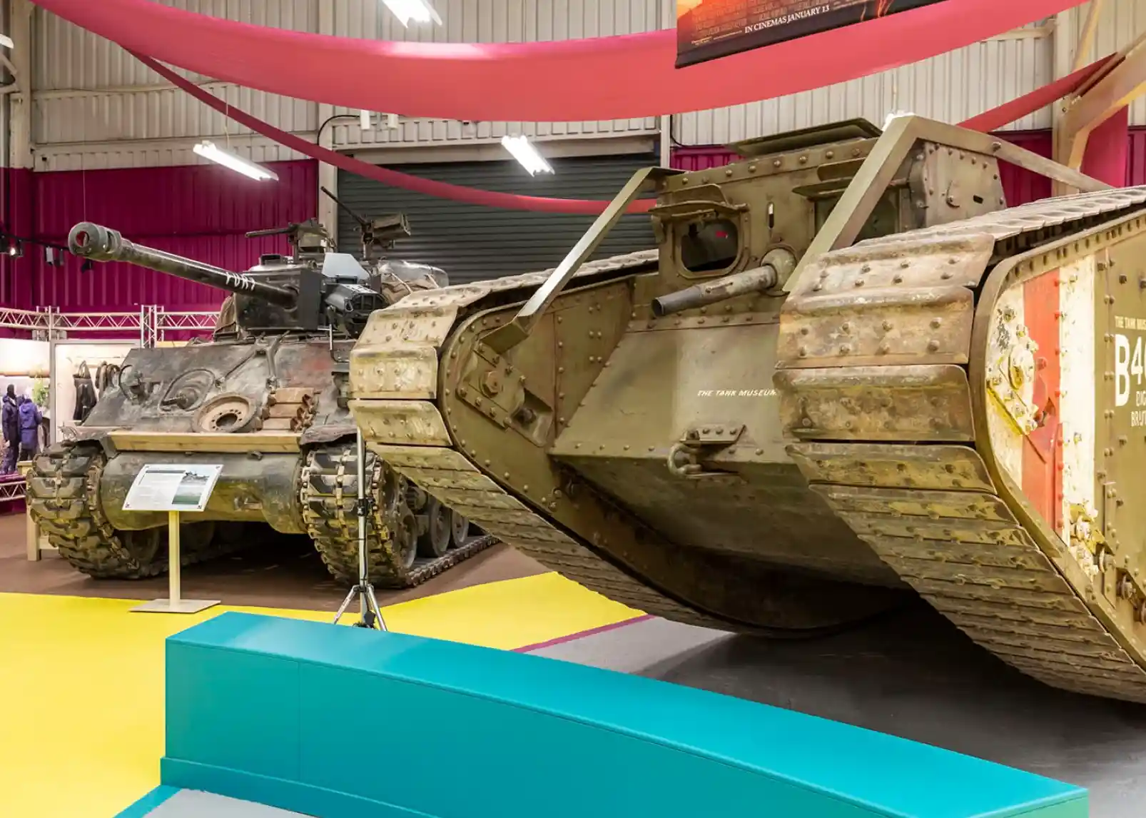 Two military tanks displayed in a museum setting. A larger, vintage tank is angled in the foreground, while a more modern tank is positioned in the background. The exhibit features colorful walls and a vibrant floor.