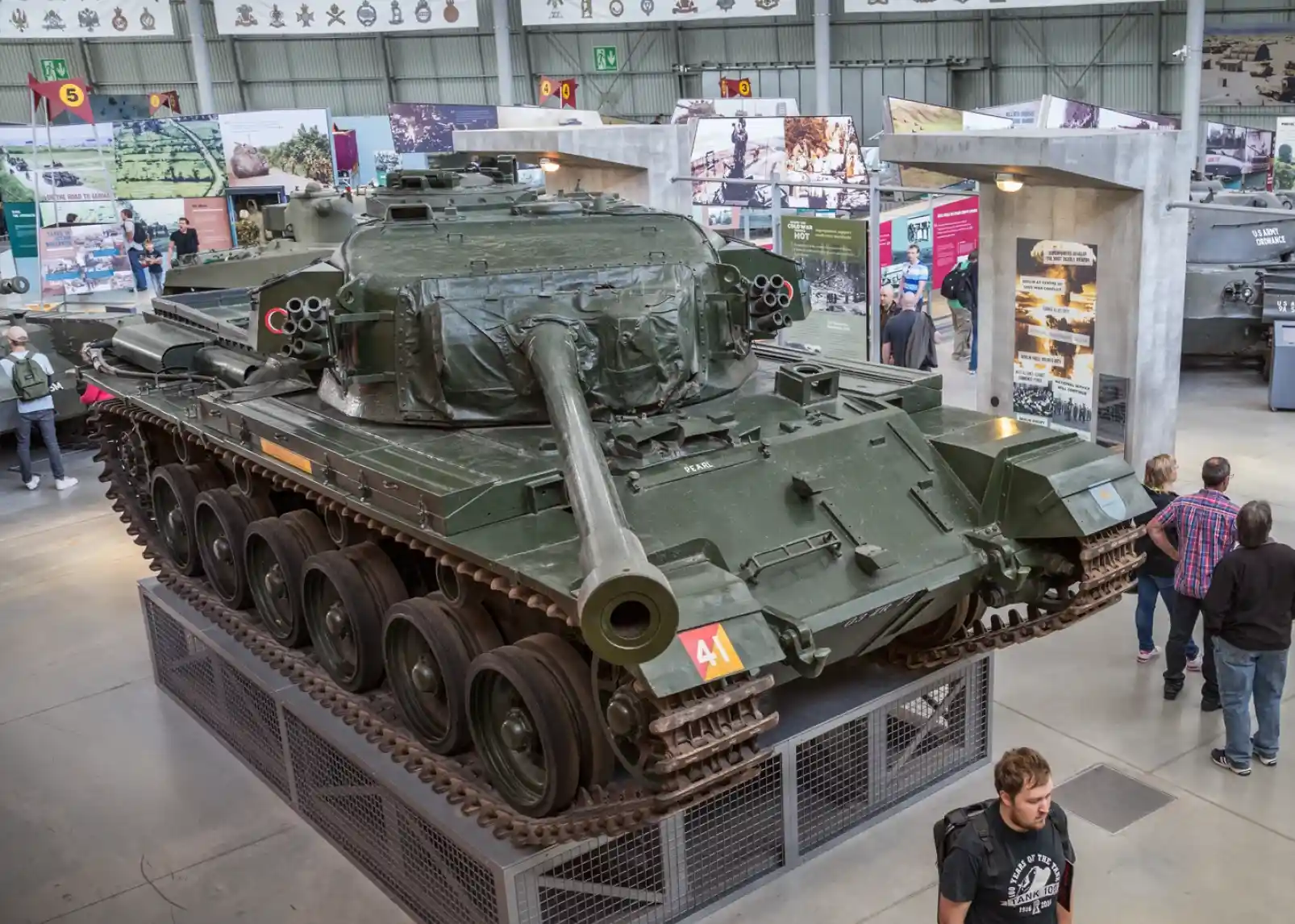 A T-55 tank is displayed prominently in an exhibition hall, surrounded by informational panels and visitors. The tank is painted in military green with a long barrel and tracks visible at the base.