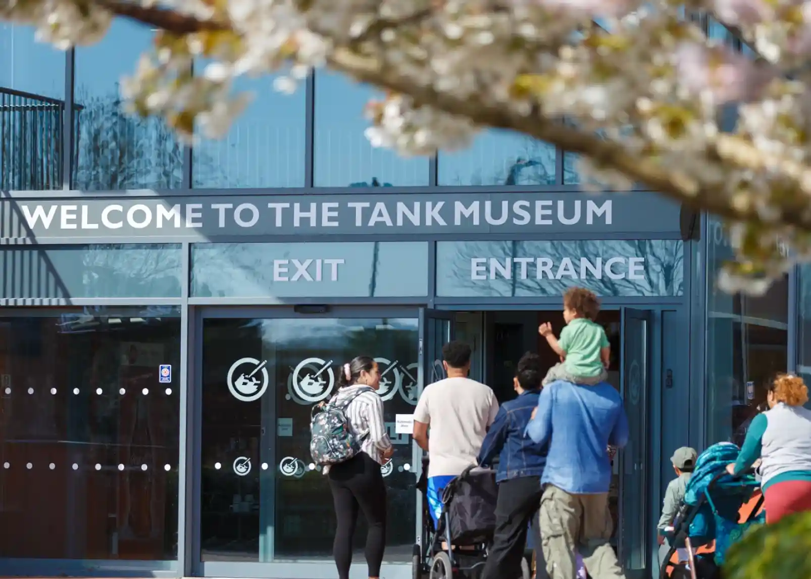 Visitors approach the entrance of the Tank Museum, with a sign that reads "Welcome to the Tank Museum." Cherry blossoms are visible in the foreground, adding a touch of spring.