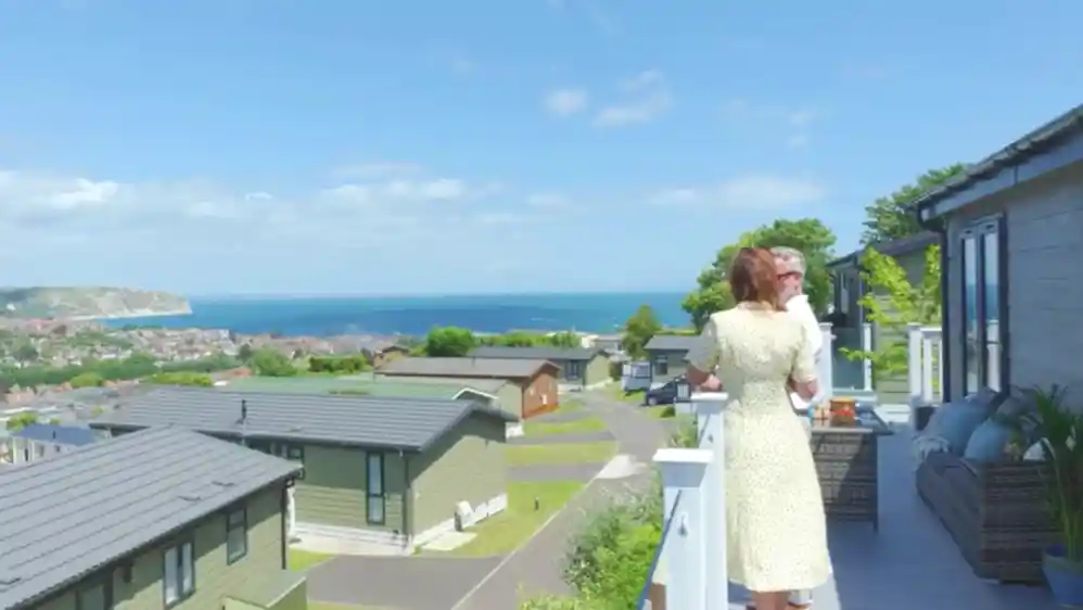 A woman in a white dress stands on a balcony, enjoying a scenic view of the ocean and coastal landscape. Nearby buildings are arranged along winding roads, with lush greenery surrounding the area under a clear blue sky.