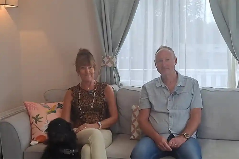 A woman and a man sit on a couch in a bright room. The woman has long hair and wears a patterned top. The man is in a light blue shirt. A black dog is sitting beside them.
