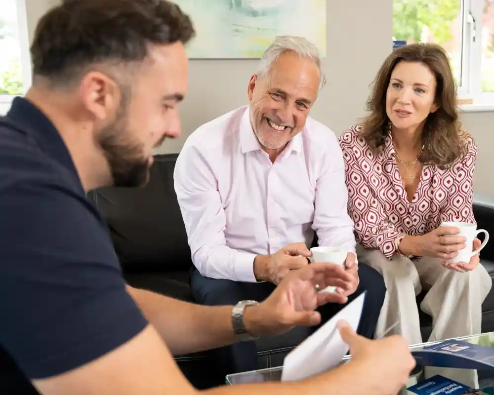 A man is smiling while sitting on a couch with a woman, both holding cups. They are engaged in conversation with another man, who is looking down at a document in his hands. The setting is bright and comfortable, with a modern decor.