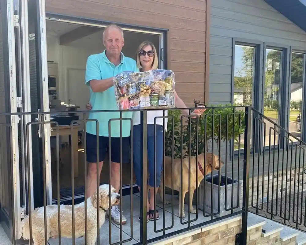 A man and a woman stand on a porch, holding a large tray filled with assorted snacks. Two dogs, one white and fluffy and the other a golden brown, are sitting beside them. The house's modern exterior is visible in the background.