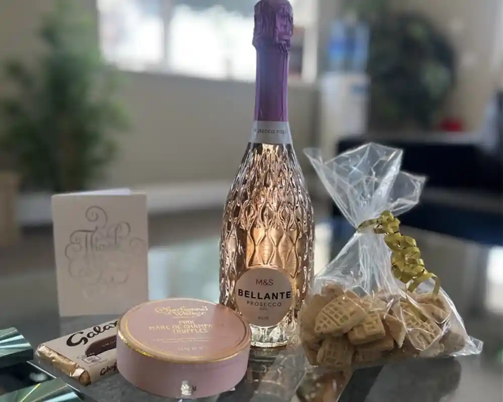 A decorative setup featuring a bottle of Bellante Prosecco with a purple cap, a box of Marc de Champagne truffles, a bag of small cookies tied with a ribbon, and a thank you card. The items are placed on a reflective table surface, with soft lighting in the background.