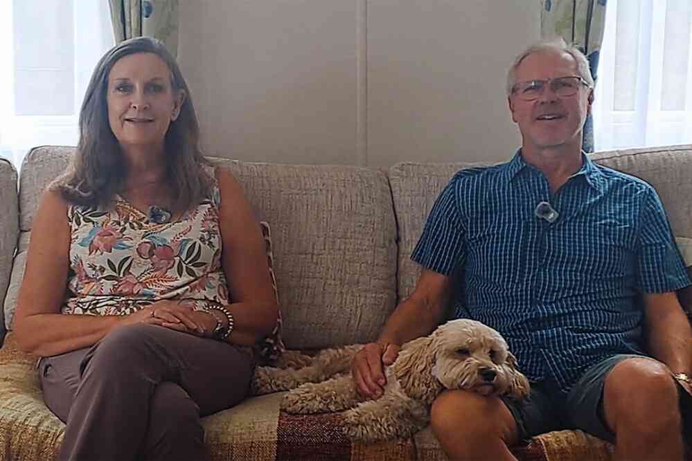 A man and a woman sit on a couch, smiling at the camera. The woman wears a floral top, and the man is in a blue checkered shirt. A small dog rests comfortably between them on the sofa.
