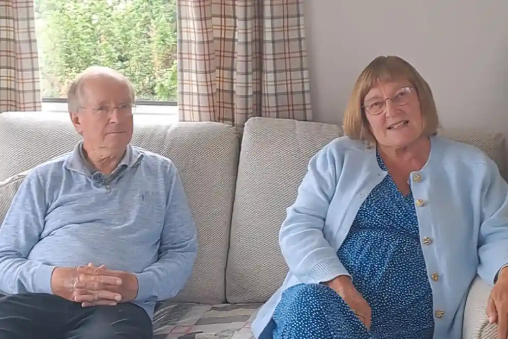 An older man and woman sit together on a sofa in a cozy living room. The man, wearing a light blue sweater, has short grey hair and looks thoughtful. The woman, dressed in a blue dress with a light blue cardigan, smiles warmly while speaking. Natural light flows in through a window, and patterned curtains frame the view of green foliage outside.