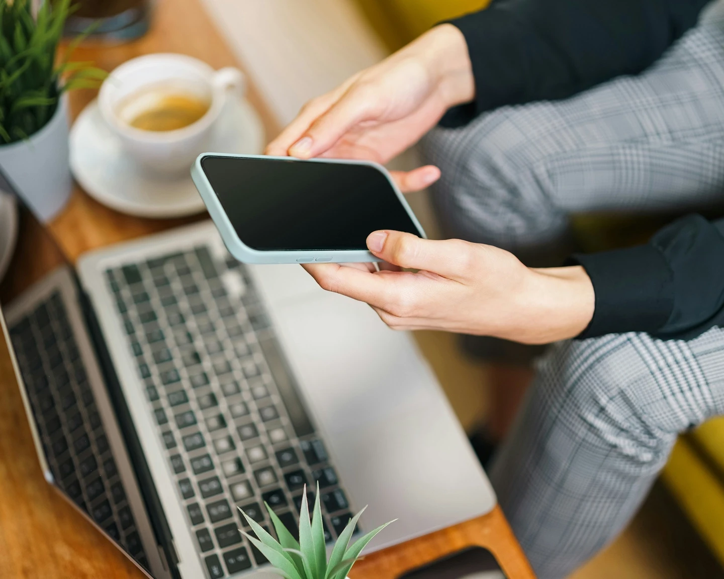 A person is holding a mobile phone looking at the blank screen with a laptop underneath on a coffee table next to a cup of tea and plants.